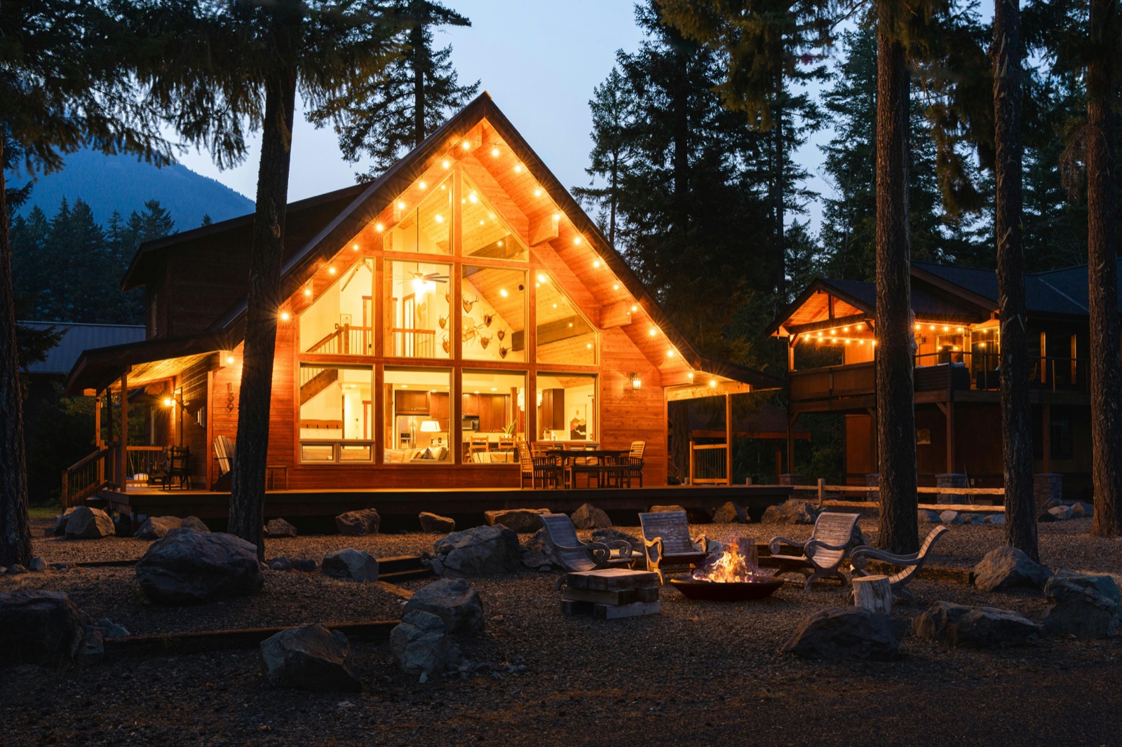 A fire pit burning beside a warmly lit cabin at dusk, string lights tracing the gable